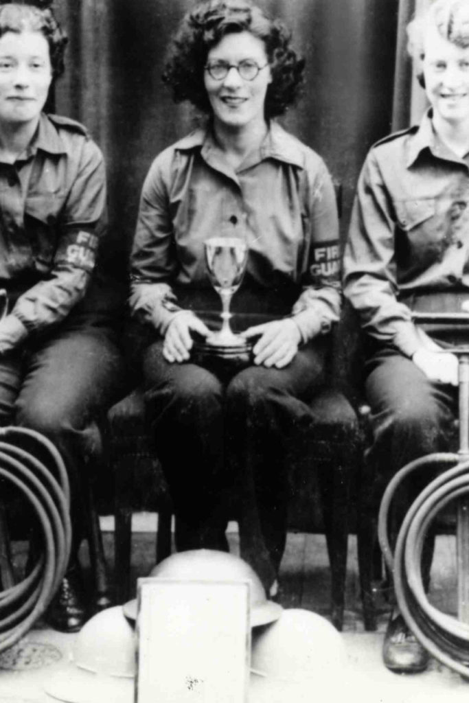 A black and white image of 3 women seated, holding a trophy and fire hoses.