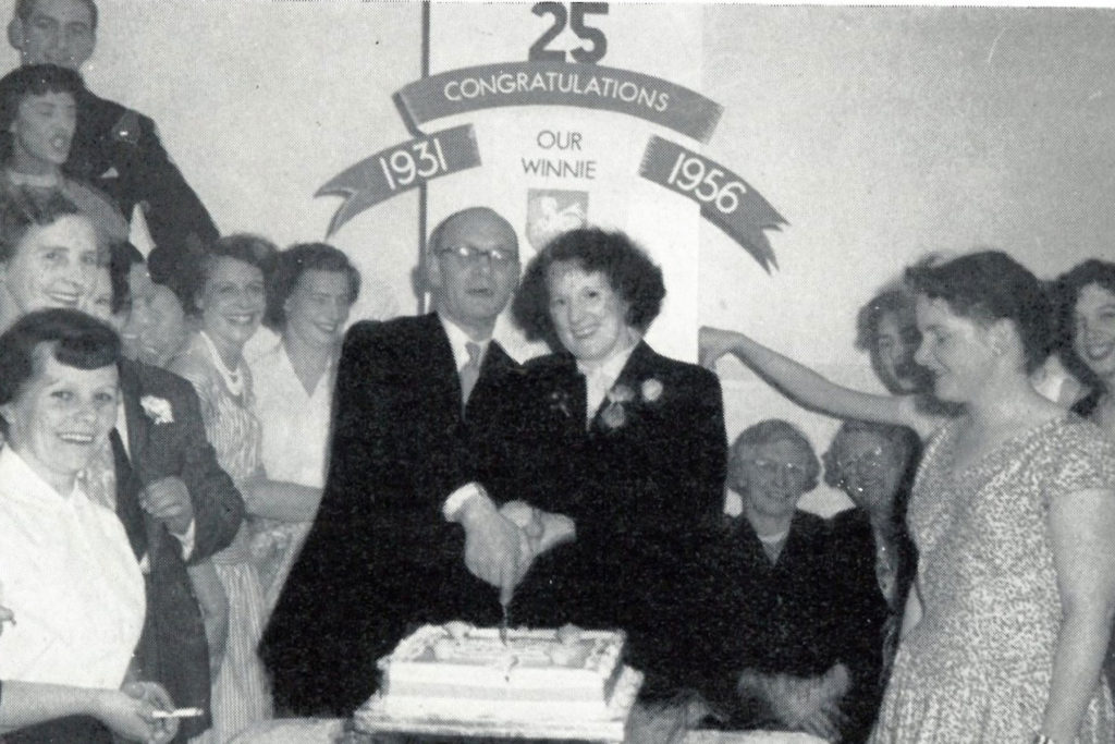A black and white image of two people cutting a big cake with a group of people celebrating.