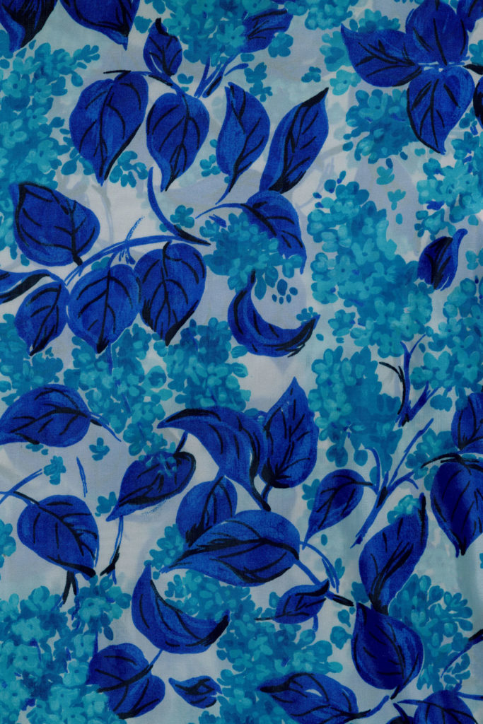A colour image of a printed fabric with a blue foliage design of royal blue leaves and teal blue small flowers on a white background.