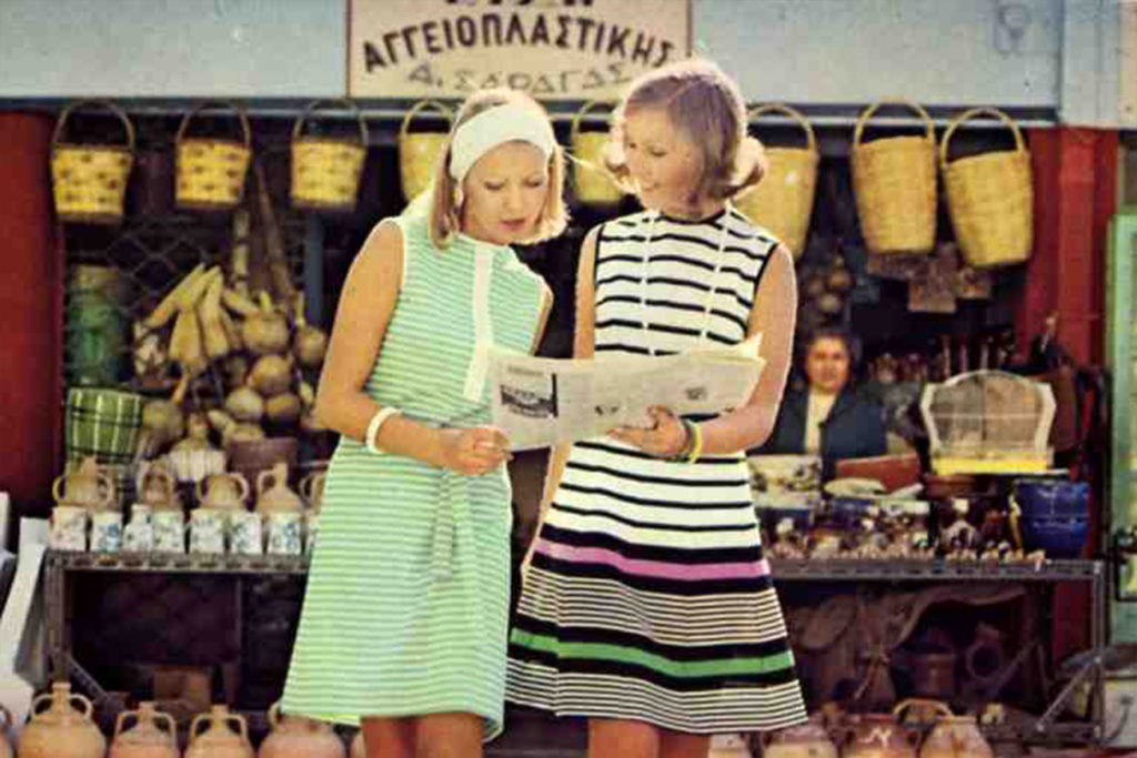 A colour image of two female models looking at a newspaper, both wear striped dresses.