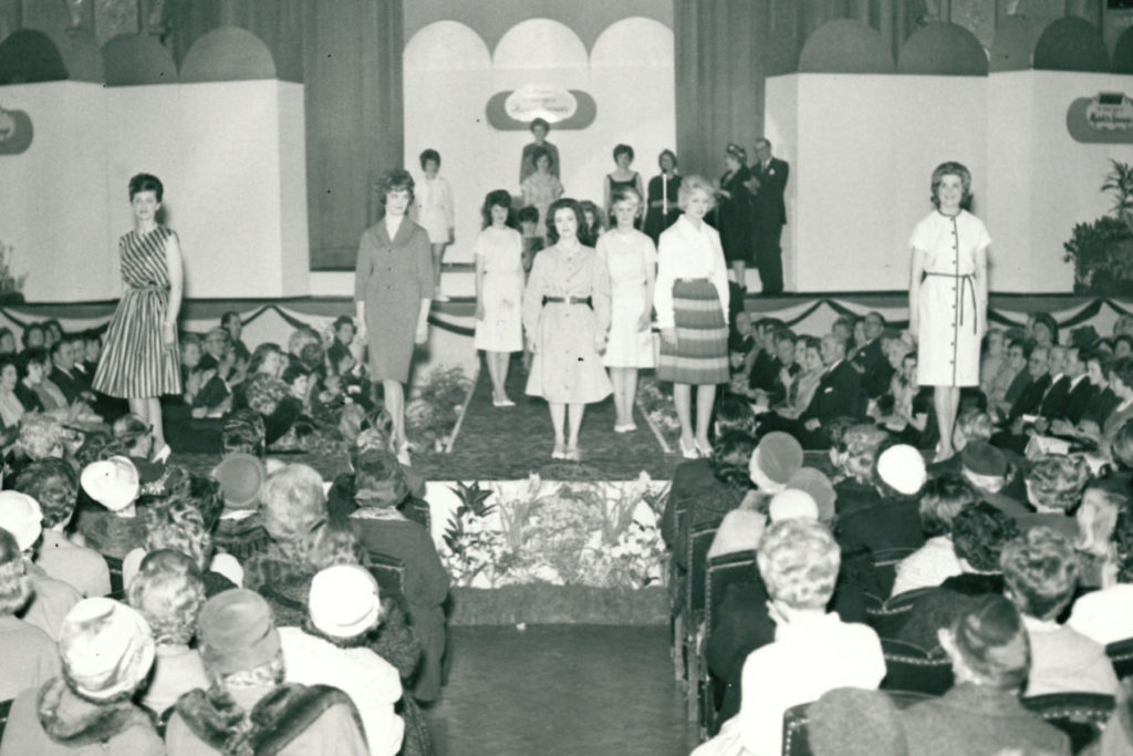Black and white image of a fashion show with models on stage and audience seated in front