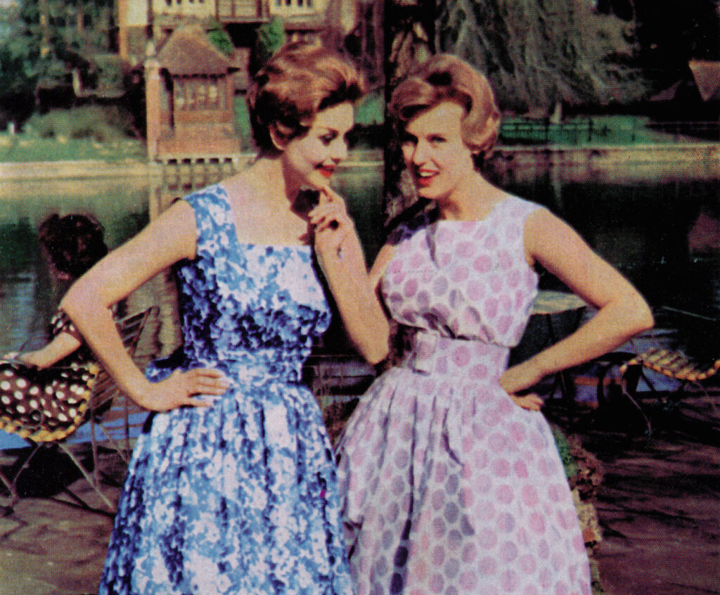 Two women stand in front of a pond, they both wear pastel coloured dresses