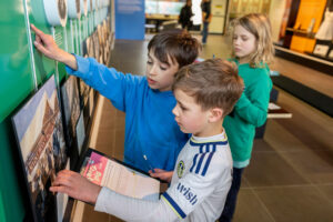 colour image of three children looking at a wall display with clipboards