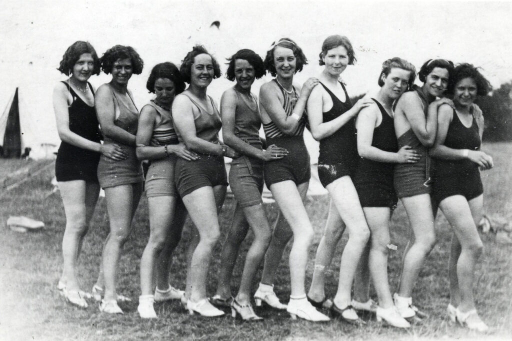 Black and white image of a group of young women standing in a line, they are outside and all are wearing swimwear.