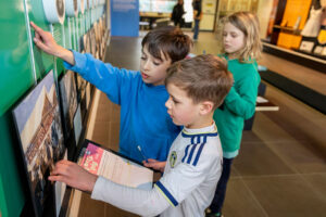 colour image of three children looking at a wall display with clipboards