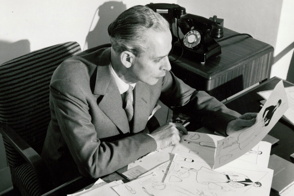 A black and white image of a man sitting at a desk looking at fashion designs.