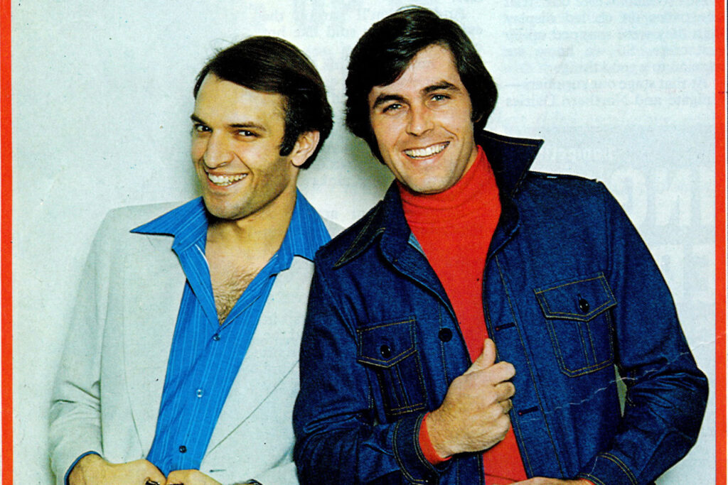 A colour image of two models, left model wears a cream jacket and blue striped shirt with wide collar, right model wears a denim jacket and red turtle neck.