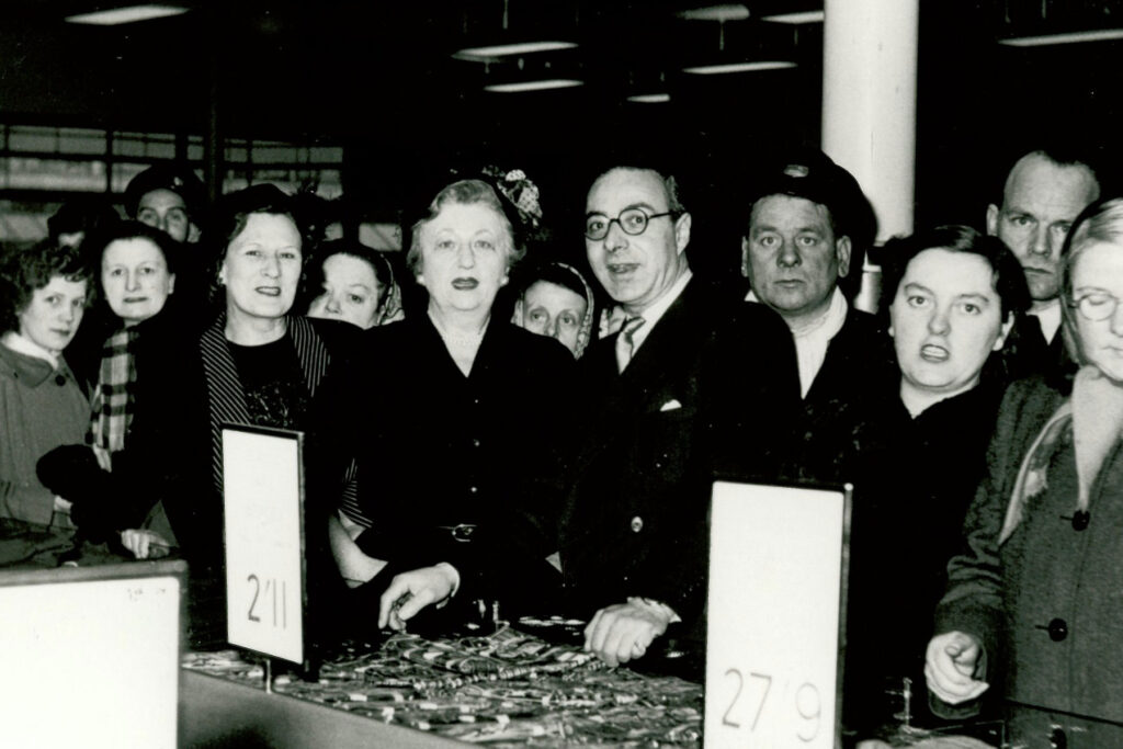 A black and white photo of a group of people all looking at the camera. They are standing in front of a shop counter.